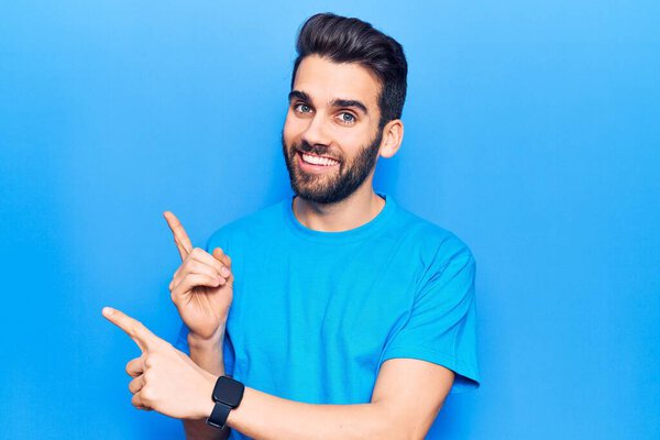 Young handsome man with beard wearing casual t-shirt smiling and looking at the camera pointing with two hands and fingers to the side. 