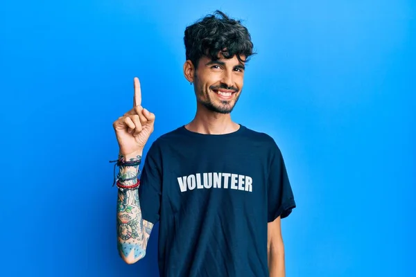ボランティアのTシャツを着た若いヒスパニック系の男性が成功したアイデアで指を指しています 興奮して幸せよ 第1位 — ストック写真