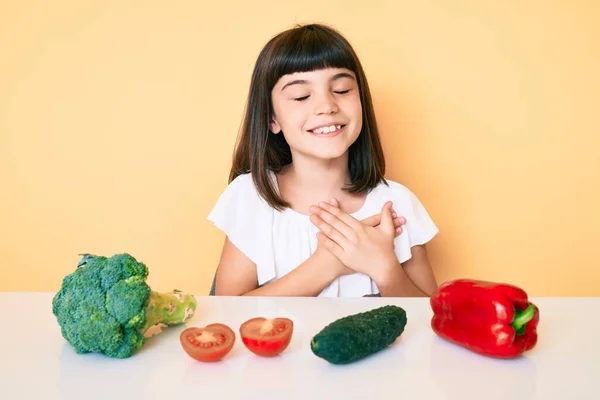 胸に手で笑みを浮かべて野菜とテーブルの上に座ってバンバンと若い女の子は 目の顔に感謝のジェスチャーで閉じます 健康の概念 — ストック写真