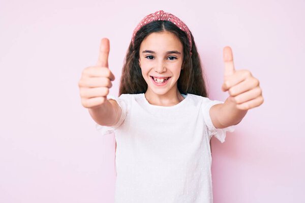 Cute hispanic child girl wearing casual clothes and diadem approving doing positive gesture with hand, thumbs up smiling and happy for success. winner gesture. 