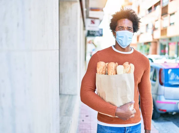 若いハンサムなアフリカ系アメリカ人の男性は医療マスクを着ている 町並みに立ってパンと紙袋を保持 — ストック写真