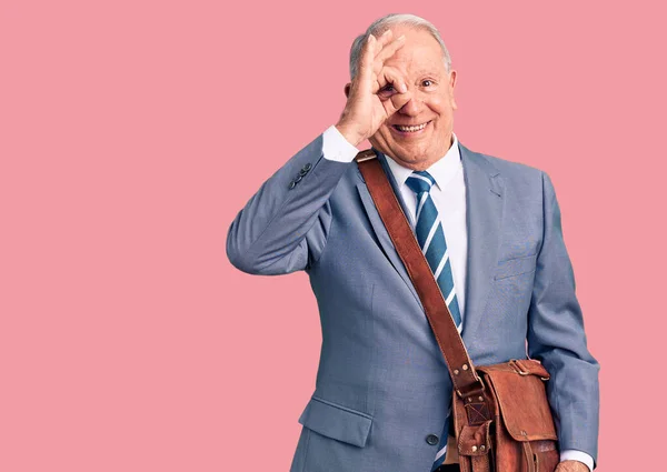 Senior handsome grey-haired man wearing suit and briefcase smiling happy doing ok sign with hand on eye looking through fingers