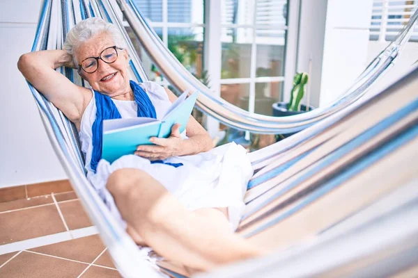 Elder senior woman with grey hair smiling happy relaxing on a hammock reading a book at home