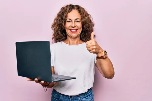 中年漂亮的女商人用笔记本电脑在孤立的粉红背景下工作时 面带微笑 面带微笑 大拇指向上做得出色 并签了字 — 图库照片