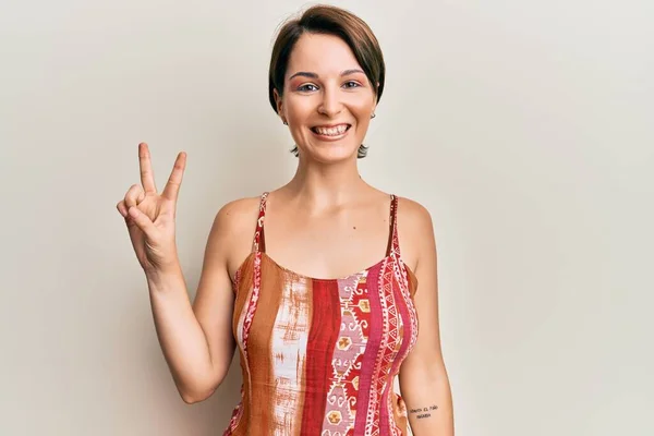 Young brunette woman with short hair wearing summer dress smiling with happy face winking at the camera doing victory sign. number two.