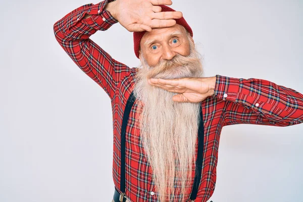 Old senior man with grey hair and long beard wearing hipster look with wool cap smiling cheerful playing peek a boo with hands showing face. surprised and exited