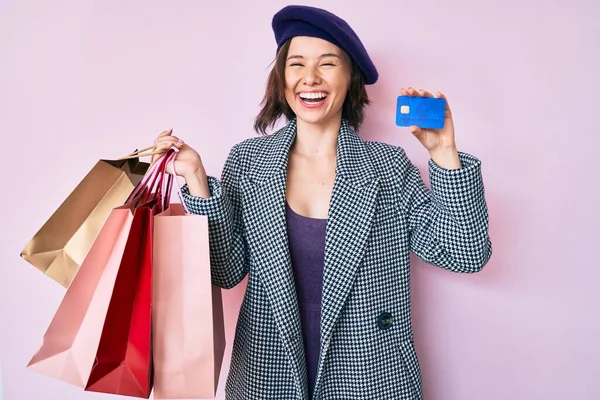 Young beautiful woman wearing beret holding shopping bags and credit card smiling and laughing hard out loud because funny crazy joke.