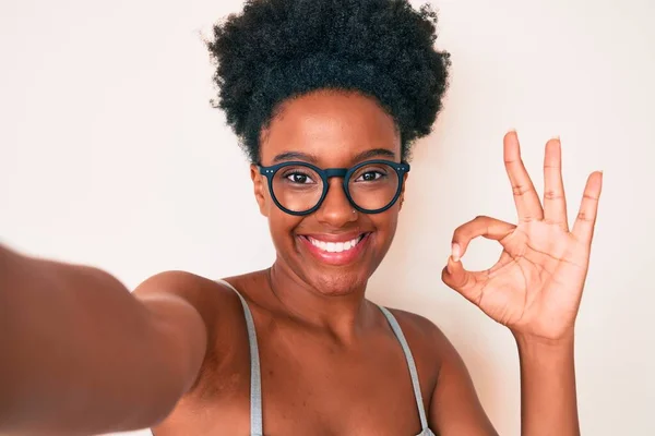 Young african american woman wearing casual clothes and glasses making selfie by the camera doing ok sign with fingers, smiling friendly gesturing excellent symbol