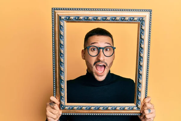 Handsome man with tattoos holding empty frame celebrating crazy and amazed for success with open eyes screaming excited.