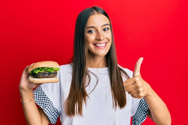 Jovem Hispânica Comendo Hambúrguer Sorrindo Feliz Positivo Polegar Para Cima — Fotografia de Stock