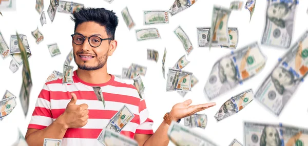 Yakışıklı Latin Amerikalı genç adam, günlük kıyafetler ve gözlük takıyor, avuç içi gösteriyor ve baş parmaklarıyla hareket ediyor, mutlu ve neşeli gülümsüyor.