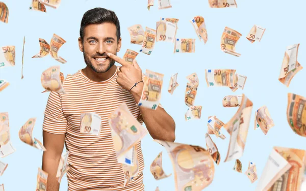 Yakışıklı İspanyol bir adam, günlük kıyafetler giyip, el parmağı ve burunla işaret ediyor, neşeli gülümsüyor. güzellik konsepti