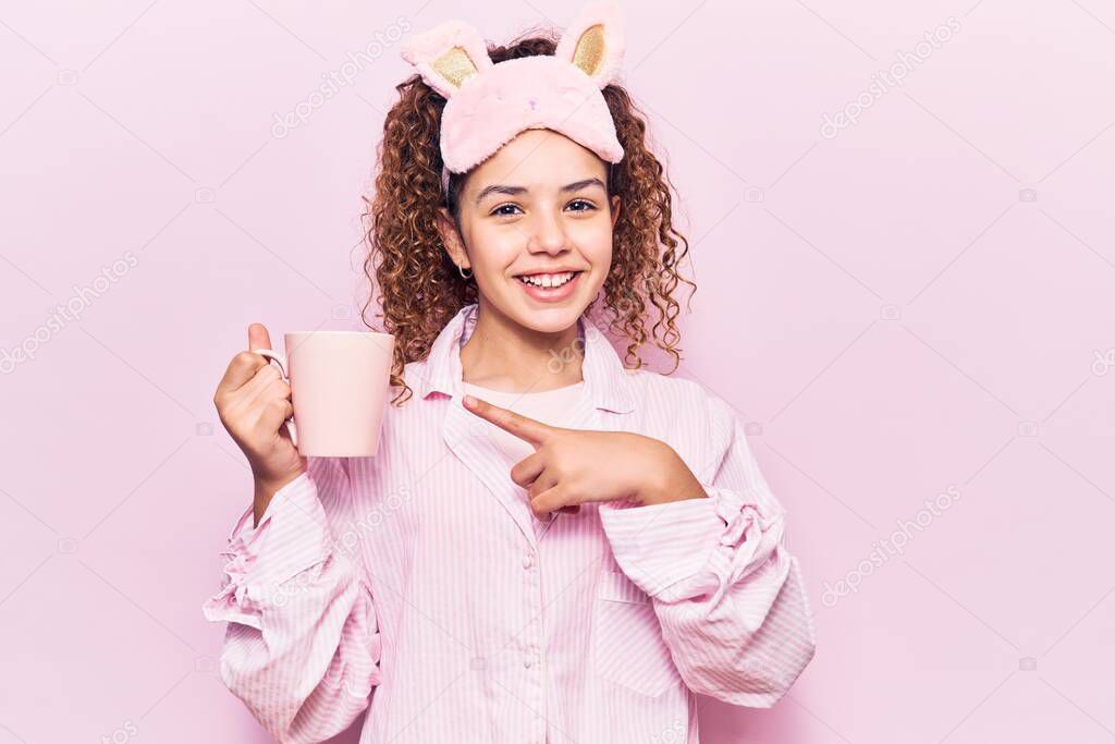 Beautiful kid girl with curly hair wearing sleep mask and pajamas holding coffee smiling happy pointing with hand and finger 