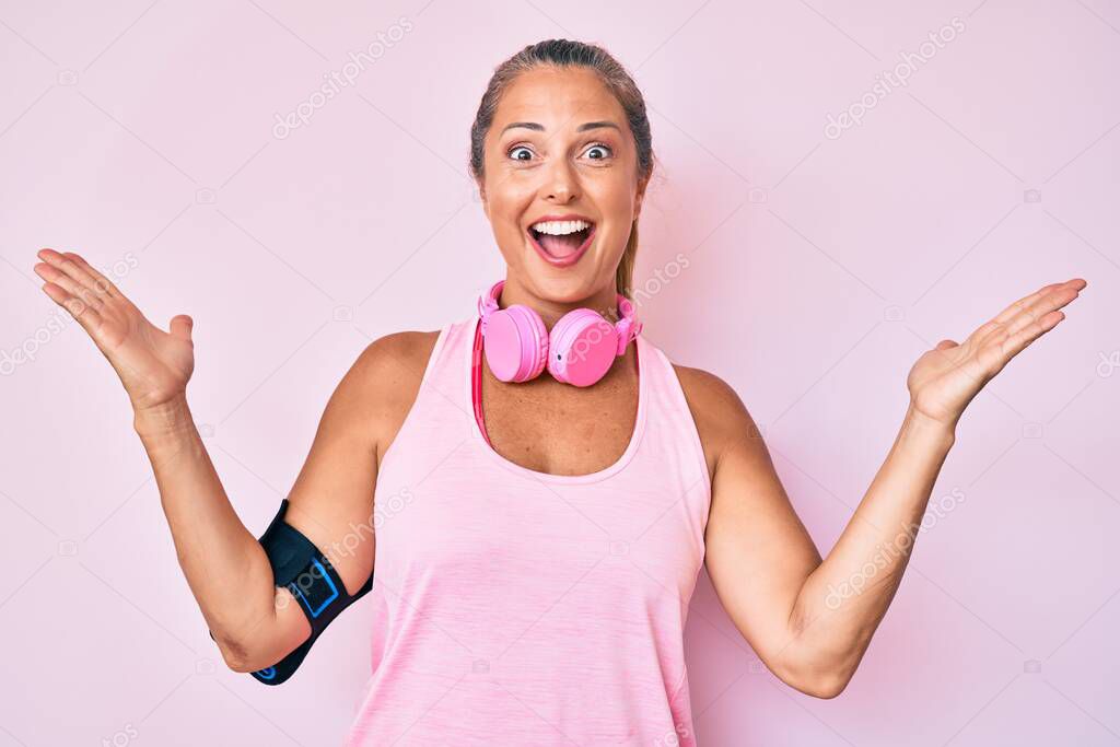 Middle age hispanic woman wearing gym clothes and using headphones celebrating victory with happy smile and winner expression with raised hands 