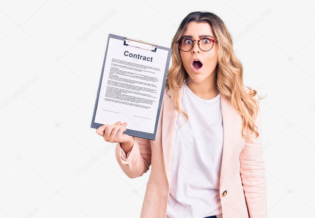 Young caucasian woman holding clipboard with contract document scared and amazed with open mouth for surprise, disbelief face 