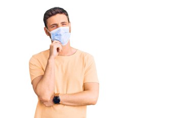 Koronavirüs için tıbbi maske takan yakışıklı genç adam. Ciddi bir yüz ifadesi. Elleri çenesinde soru sormayı düşünen, kafa karıştırıcı bir fikir. 