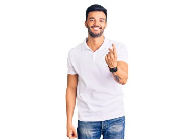 Genç, yakışıklı, günlük kıyafetler giymiş İspanyol bir adam, el kol hareketi yaparak, mutlu ve gülümseyen birini davet ediyor. 