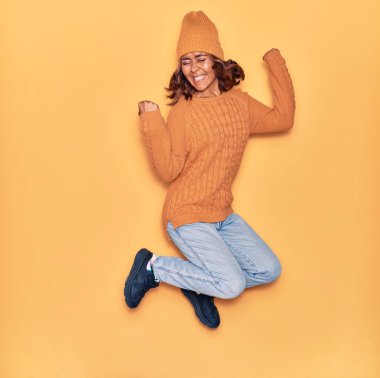 Yün şapka ve süveter giyen, mutlu gülümseyen genç, güzel Latin bir kadın. Yüzünde tebessümle zıplıyor, sarı arka planda yumruklar havada kutluyor.