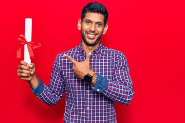 Diplomasını elinde tutan genç bir latin adam mutlu bir şekilde elini ve parmağını göstererek gülümsüyor. 