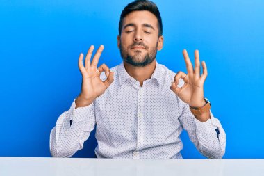 İş kıyafetleri giymiş yakışıklı İspanyol bir adam masaya rahatça oturuyor ve gözleri kapalı gülümsüyor parmaklarıyla meditasyon hareketi yapıyor. yoga konsepti. 