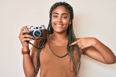 Saçları örgü örmüş, elinde antika bir kamerayla gülümseyen mutlu ve gururlu bir kadını işaret eden Afrikalı genç bir Amerikalı kadın. 