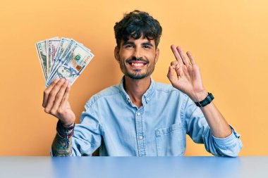 Masada dolarları tutan İspanyol genç adam parmaklarıyla imza atıyor, gülümseyerek mükemmel bir sembol oluşturuyor. 