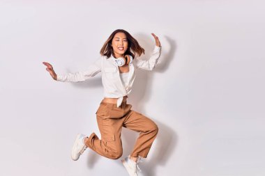 Genç, güzel Çinli kadın mutlu gülümsüyor. Yüzünde gülümsemeyle zıplıyor, beyaz arka planda kulaklık kullanarak müzik dinliyor.
