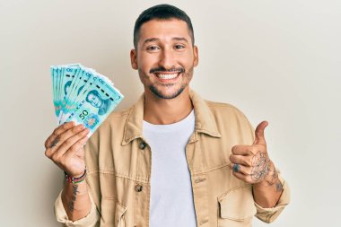 50 Yuan 'lık Çin banknotları tutan yakışıklı adam mutlu ve pozitif gülümsüyor, başparmağıyla mükemmel ve onay işareti yapıyor. 