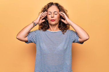 Orta yaşlı, çizgili tişört giyen güzel bir kadın izole edilmiş sarı arka planda duruyor. Başında baş ağrısı var çünkü stres var. Migren ağrısı..