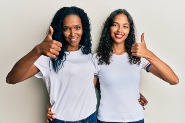 Afro-Amerikan anne ve kızı günlük beyaz tişört giyiyorlar mutlu ve pozitif gülümsüyorlar, başparmağıyla mükemmel ve onay işareti yapıyorlar. 