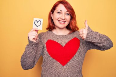 Güzel, kızıl saçlı bir kadın, sarı arka planda kalbi olan mutlu ve mutlu bir şekilde gülümseyen, başparmağını kaldırarak mükemmel ve onay işareti yapan bir kadın.