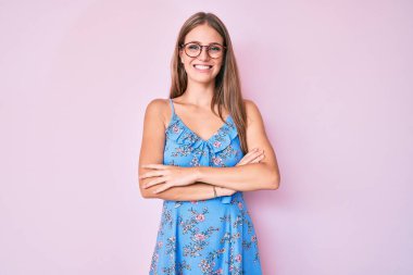 Sıradan kıyafetler giyen, gözlüklü mutlu yüzlü, kolları çapraz bir şekilde kameraya bakan genç sarışın kız. pozitif kişi. 