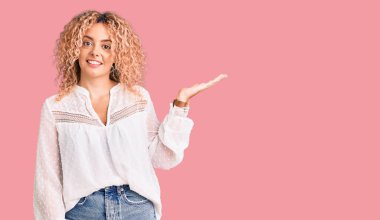 Kıvırcık saçlı, zarif bir yaz gömleği giyen neşeli bir şekilde gülümseyen ve avuç içi kameraya bakan genç sarışın kadın.. 