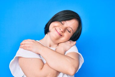 Down sendromlu esmer bir kadın, günlük kıyafetler giyiyor, kendine sarılıyor, mutlu ve pozitif, kendinden emin gülümsüyor. Sevgi ve özgüven. 