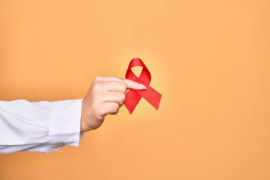 İzole edilmiş sarı arka plan üzerinde HIV hastalığını destekleyen kırmızı kanser kurdelesi tutan beyaz bir kadının eli.