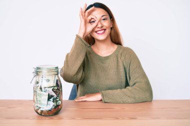 Masada oturan genç esmer kadın. Dolarla dolu kavanozu var. Gülümsüyor. 