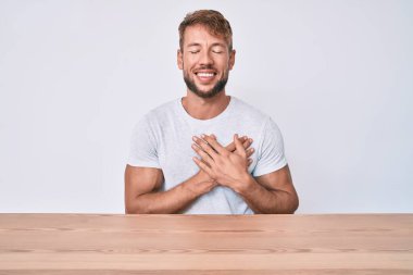 Gündelik giysiler giymiş genç beyaz bir adam masaya oturmuş elleri göğsünde, gözleri kapalı ve yüzünde minnet dolu bir jest ile oturuyor. sağlık kavramı. 