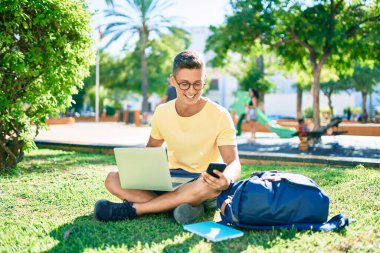 Üniversite kampüsünde çimlerin üzerinde otururken mutlu bir şekilde gülümseyen İspanyol öğrenci.