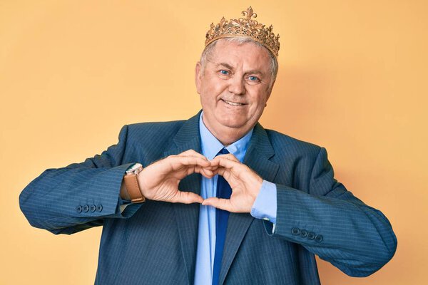 Старший седовласый мужчина в деловом костюме и королевской короне, улыбающийся в любви, показывает символы и форму сердца руками. романтическая концепция. 