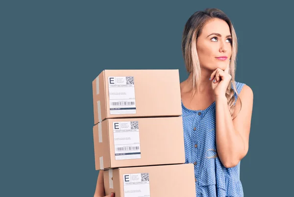 Young Beautiful Blonde Woman Holding Delivery Boxes Serious Face Thinking — Stock Photo, Image