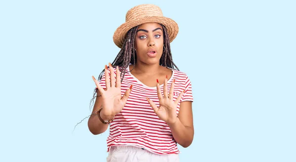 Young african american woman with braids wearing summer hat moving away hands palms showing refusal and denial with afraid and disgusting expression. stop and forbidden.