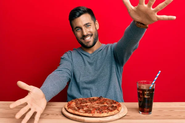 抱擁のために腕を開いて笑顔カメラを見ておいしいペパロニピザを食べるハンサムなヒスパニック系の男 幸せを受け入れる陽気な表情 — ストック写真