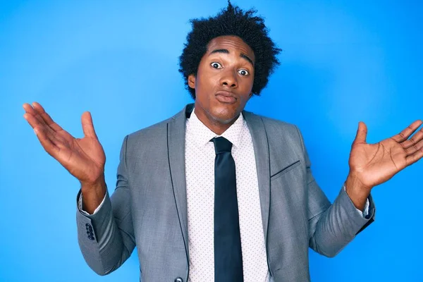 Afro saçlı, iş ceketinden bihaber ve kolları açık bir şekilde kafası karışmış yakışıklı Afro-Amerikalı bir adam.. 