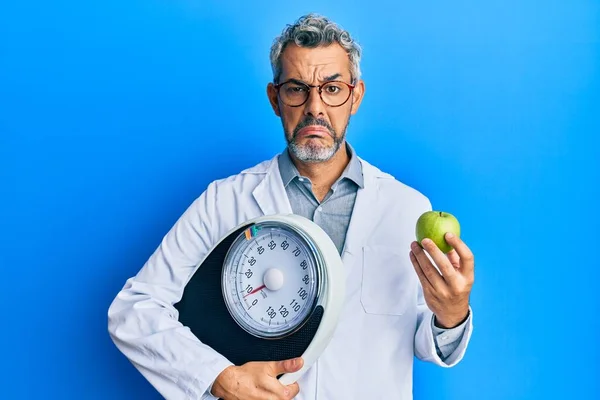 中年白发男子作为营养师医生 拿着称重机和绿色苹果 忧心忡忡 怒气冲冲 胆战心惊 悲伤的表情 — 图库照片