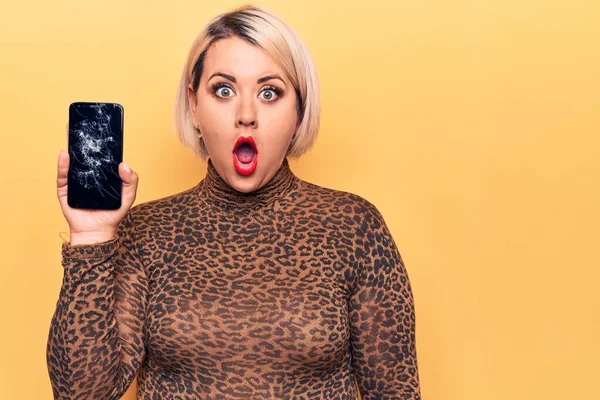 Young beautiful blonde plus size woman holding broken smartphone showing cracked screen scared and amazed with open mouth for surprise, disbelief face