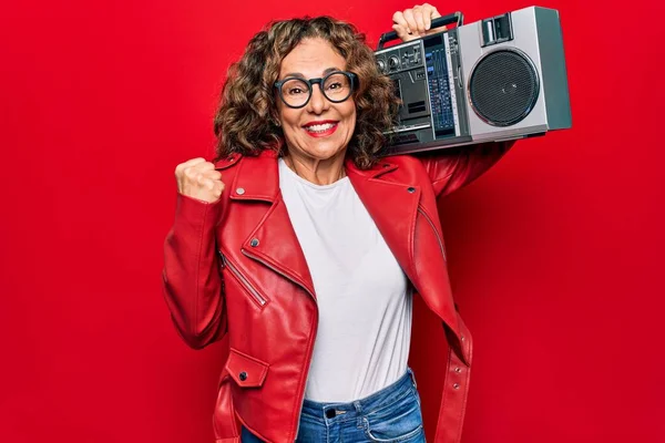 Middle age brunette hipster woman holding retro music boombox over red isolated background screaming proud, celebrating victory and success very excited with raised arm