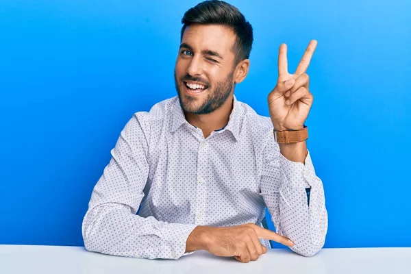 ビジネス用の服を着たハンサムなヒスパニック系の男性がテーブルの上に座って カメラで勝利のサインを指で行う幸せな顔をウィンクで笑っています — ストック写真