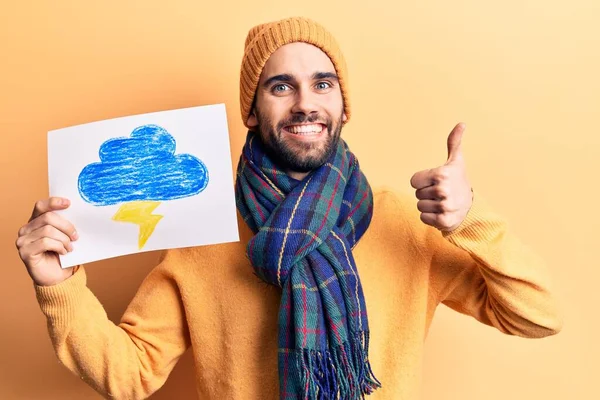 Young handsome man with beard wearing winter clothes holding thunder draw smiling happy and positive, thumb up doing excellent and approval sign