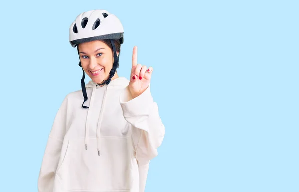 Beautiful brunette young woman wearing bike helmet and sporty clothes showing and pointing up with finger number one while smiling confident and happy.