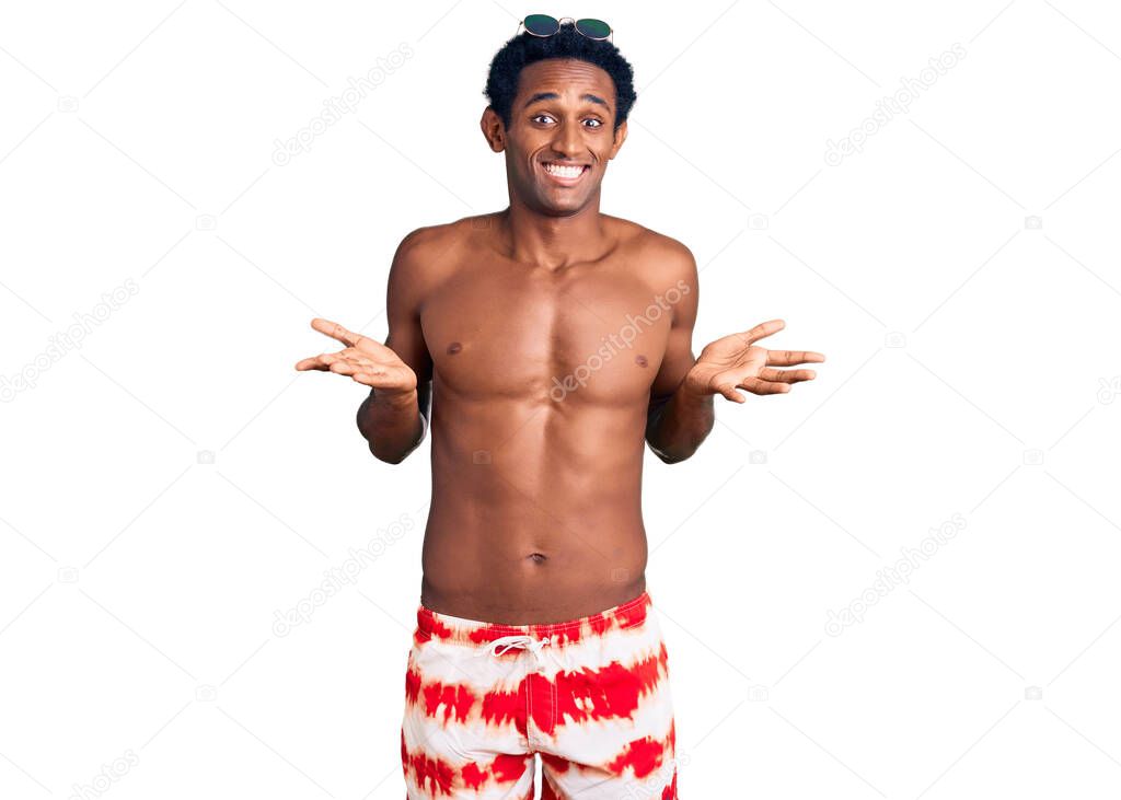 African handsome man wearing swimsuit and sunglasses clueless and confused expression with arms and hands raised. doubt concept. 